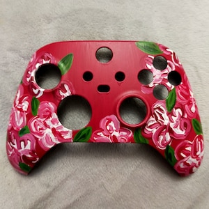 Puede incluir: Un mando de videojuegos rojo con un diseño floral pintado en él. Las flores son rosas y blancas con hojas verdes.