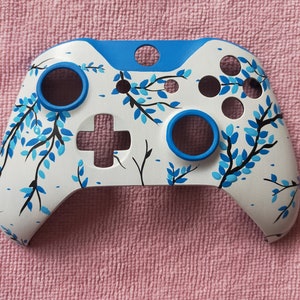Puede incluir: Un mando de videojuego personalizado pintado en blanco y azul con un diseño floral. El mando tiene un acento azul alrededor de los bordes y los botones.