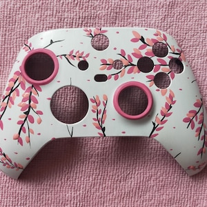 Pode incluir: Controlador de console de jogos branco com design floral rosa e detalhes em rosa. O controlador tem dois botões rosas e dois gatilhos rosas.