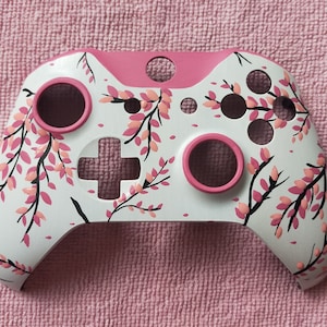 May include: A white Xbox controller with a pink and black floral design. The controller has a pink faceplate and pink buttons.