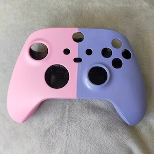 May include: A pink and purple video game controller shell. The shell is split down the middle, with the left side being pink and the right side being purple. The shell is missing the buttons and joysticks.