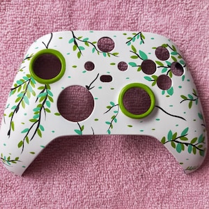 Puede incluir: Un mando Xbox blanco con un diseño de hojas verdes y negras. El mando tiene un botón verde en la parte delantera.