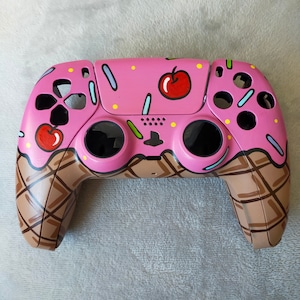 May include: A pink and brown custom-painted video game controller with a waffle cone and ice cream design. The controller has a pink base with brown waffle cone accents and sprinkles. There are two red cherries on the top of the controller.