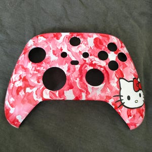 Puede incluir: Un mando de videojuegos personalizado con un diseño veteado en rojo, rosa y blanco. El mando tiene botones negros y un gráfico de Hello Kitty en el lado derecho. El fondo es una tela gris oscuro.
