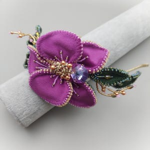 May include: A handmade purple flower brooch, featuring a central cluster of gold beads and a large, faceted, light purple gem. The flower's petals are edged with small gold beads. Green beaded leaves and gold wire accents complete the design.