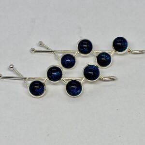 May include: Two silver hair pins, each with a series of dark blue, spherical beads. The beads have a speckled, galaxy-like appearance. The pins are arranged side-by-side against a white background.