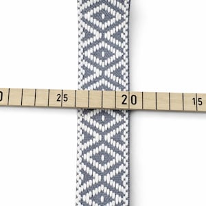 Könnte beinhalten: Ein graues und weißes gewebtes Uhrenarmband mit einem sich wiederholenden Rautenmuster. Ein Holzlineal liegt horizontal über dem Band und zeigt eine Breite von etwa 2,2 Zentimetern an der Stelle des Kontakts mit dem Lineal.
