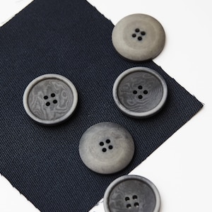 Könnte beinhalten: Fünf graue Knöpfe mit jeweils vier Löchern. Die Knöpfe liegen auf einem dunkelblauen Stoff.