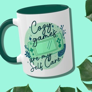 May include: White ceramic mug with a green rim and handle. The mug features a green and blue illustration of a handheld video game console with the text "Cozy games are my self care" in a stylized font.