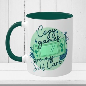May include: White ceramic mug with a green rim and handle. The mug features a green and blue illustration of a video game controller with the text "Cozy games are my self care" in a circle.