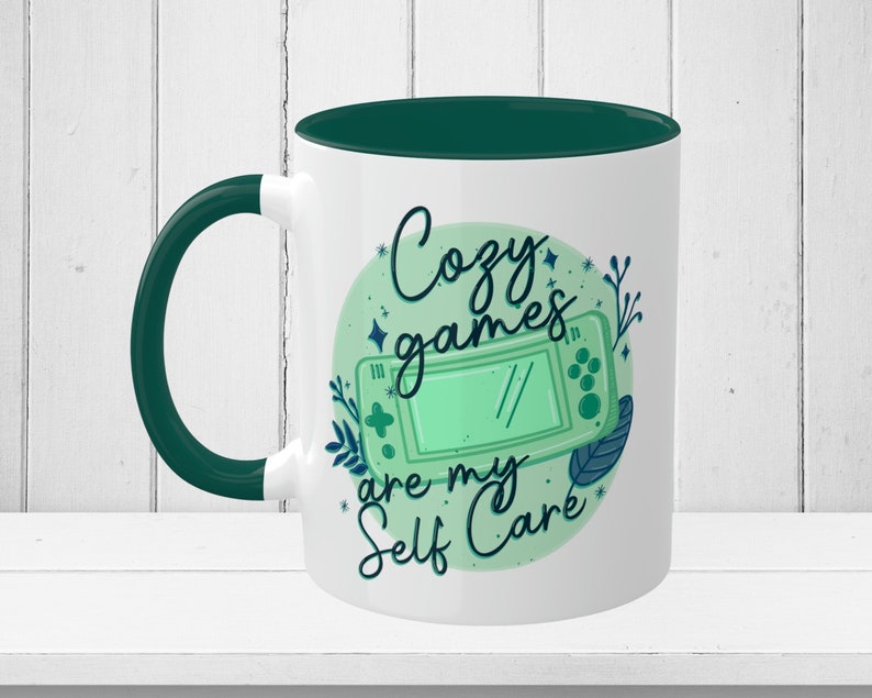 May include: White ceramic mug with a green rim and handle. The mug features a green and blue illustration of a video game controller with the text "Cozy games are my self care" in a circle.