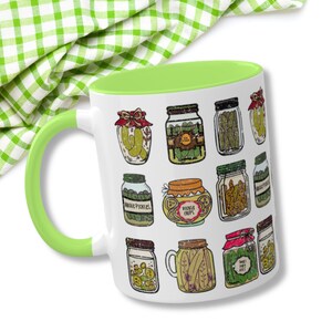 K&ouml;nnte beinhalten: Eine wei&szlig;e Keramiktasse mit gr&uuml;nem Rand zeigt eine farbenfrohe Illustration verschiedener Gl&auml;ser, gef&uuml;llt mit Gurken, Gurkenchips und s&uuml;&szlig;en Gurken. Die Tasse ist perfekt f&uuml;r Gurkenliebhaber.