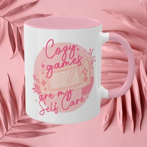 May include: A pink and white ceramic mug with a pink handle. The mug features a pink circle with a pink video game controller and the text "Cozy games are my self care" in pink.