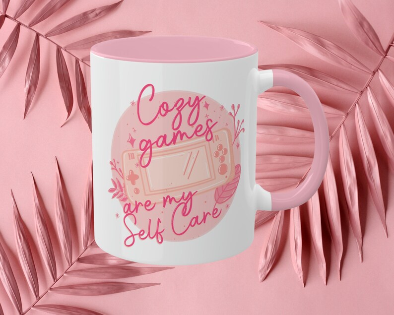 May include: A pink and white ceramic mug with a pink handle. The mug features a pink circle with a pink video game controller and the text "Cozy games are my self care" in pink.