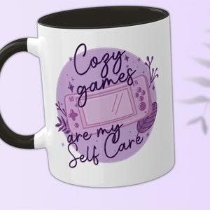 May include: White ceramic mug with a black handle and a black rim. The mug features a purple and white illustration of a video game controller with the text "Cozy games are my self care" in purple.