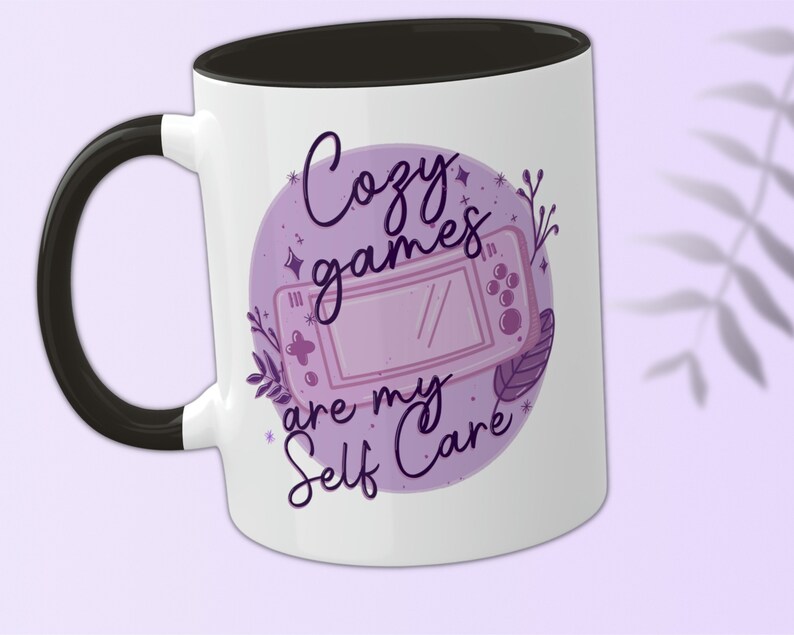 May include: White ceramic mug with a black handle and a black rim. The mug features a purple and white illustration of a video game controller with the text "Cozy games are my self care" in purple.