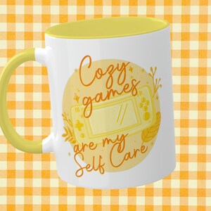 May include: White ceramic mug with a yellow rim and handle. The mug features a yellow circle with a stylised illustration of a handheld video game console and the text "Cozy games are my self care".
