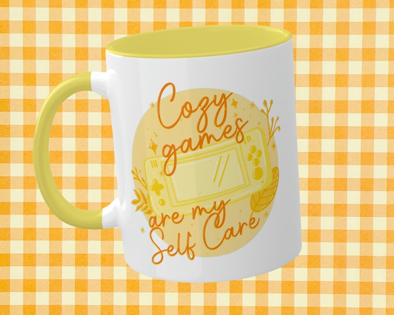 May include: White ceramic mug with a yellow rim and handle. The mug features a yellow circle with a stylised illustration of a handheld video game console and the text "Cozy games are my self care".