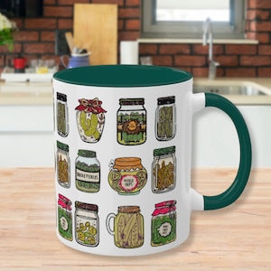 K&ouml;nnte beinhalten: Wei&szlig;e Keramiktasse mit gr&uuml;nem Rand und Henkel. Die Tasse zeigt ein Muster aus 16 illustrierten Gurkengl&auml;sern in verschiedenen Stilen. Einige Gl&auml;ser haben Etiketten mit der Aufschrift "Pickle Chips", "Sweet Baby Dills" und "Sour Pickles".