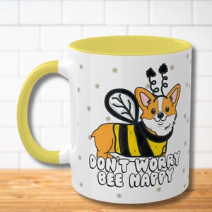 May include: White ceramic mug with a yellow interior and handle. The mug features a cartoon corgi dressed as a bee, with the text "DON'T WORRY BEE HAPPY". Small daisy illustrations are scattered across the white surface.