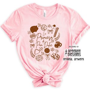 May include: A light pink t-shirt featuring a brown graphic design of various pastries and the text "La Princesa de Pan Dulce". The design includes a pig-shaped pastry, bows, and other baked goods.