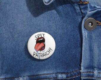 May include: A white button with a red mouth and tongue sticking out, with the text "SPIT on the PATRIARCHY" around it.