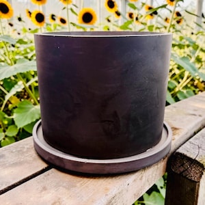May include: A dark charcoal-colored cylindrical planter with a matching saucer sits on a weathered wooden surface. The planter is empty, ready for planting. The background features sunflowers in a greenhouse setting.
