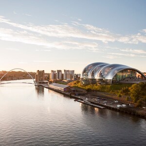 May include: A view of the Tyne River in Newcastle, England, with the Sage Gateshead concert hall and the Millennium Bridge in the background. The sun is setting over the city, casting a warm glow on the buildings.