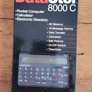 May include: A vintage DataStor 8000 C pocket computer in its original packaging. The black box features red and white text, highlighting features like 8K memory and a calculator. The device itself has a keyboard layout with an LCD screen.