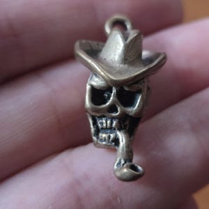 May include: A small, bronze-colored skull pendant wearing a cowboy hat and smoking a pipe. The skull has detailed features, including eye sockets, teeth, and a nose. The pendant has a loop at the top for attaching to a chain or keychain.