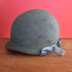 May include: An olive drab military helmet with a textured surface and a chin strap. The helmet sits on a wooden surface against a red background. The chin strap is a light green color.