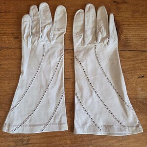 May include: A pair of white leather gloves featuring a mid-length design. Each glove is detailed with three diagonal rows of decorative stitching that run across the back of the hand and extend toward the wrist, creating a subtle pattern.