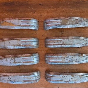May include: Eight vintage, silver-painted metal door handles with a distressed finish. The handles have a streamlined, bullet-like shape with a rounded end and a pointed tip. The paint is chipped, revealing the metal underneath. The handles are arranged on a wooden surface.