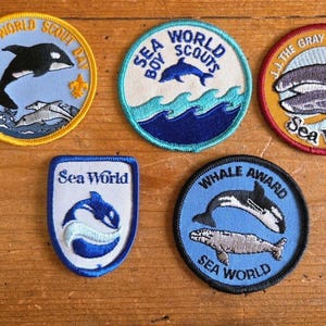 May include: A collection of Sea World patches featuring marine life. Designs include orcas, dolphins, and whales in various colors. Some patches have text like "Sea World Scouts" and "Whale Award". The patches are round and shield-shaped.