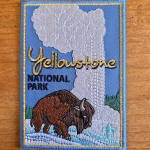 May include: Embroidered patch depicting a bison and a geyser on a blue background. The text "Yellowstone NATIONAL PARK" is in gold and navy blue. The patch has a blue border with gold stitching.