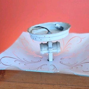 Könnte beinhalten: Vintage Deckenleuchte mit einem weißen, quadratischen Glasschirm mit einem dekorativen Blumenmuster in Kupferfarbe. Die Leuchte hat einen weißen Metallsockel und eine weiße, runde Deckenplatte.