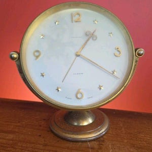Pode incluir: Um despertador vintage dourado com mostrador branco. O relógio apresenta ponteiros dourados, marcadores em forma de estrela e grandes algarismos dourados às 3, 6, 9 e 12 horas. Possui uma base redonda e dois botões laterais.