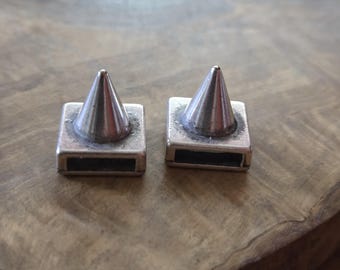 May include: Two silver-colored metal studs with a cone-shaped top and a rectangular base. The studs are displayed on a wooden surface. The studs have a dark rectangular opening on the base. The studs are likely used for clothing or accessories.
