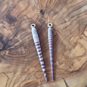 May include: Two elongated, natural-toned sea urchin spines with a striped pattern in shades of purple and white. Each spine has a silver-colored metal loop at the top, ready for jewelry making or crafting. The background is a wooden surface.