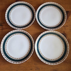 May include: A set of four round white ceramic plates, each featuring a wide dark green border decorated with a repeating gold-colored filigree pattern along the inner and outer edges of the rim, creating a consistent design across all four pieces.