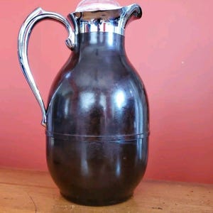 Può includere: Un thermos marrone vintage con manico, bordo e coperchio argentati. Il thermos ha una parte superiore trasparente e arrotondata e un manico curvo. Il corpo del thermos è di colore marrone scuro. Il thermos è su una superficie di legno.