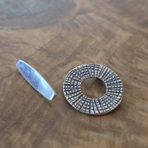 Op de afbeelding: Een zilverkleurige metalen sieradensluiting met een getextureerde, cirkelvormige hanger en een aparte, langwerpige staaf. De hanger heeft een gedetailleerd, radiaal patroon. De sluiting wordt getoond op een houten oppervlak.
