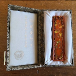 Op de afbeelding: Een open, rechthoekige doos met een decoratief patroon aan de buitenkant. Binnenin ligt een rechthoekige oranje inktstaaf met gouden accenten op witte stof. De linkerkant van de doos heeft tekst en een cirkelvormig embleem.