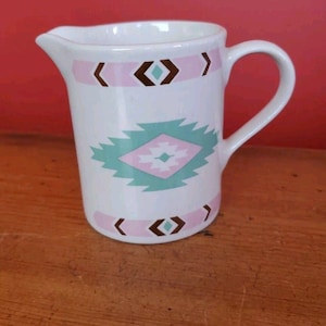Könnte beinhalten: Ein weißes Milchkännchen aus Keramik mit Henkel und Ausgießer. Die Außenseite ist mit horizontalen Bändern aus rosa und braunen geometrischen Formen sowie einem zentralen Rautenmuster in Rosa- und Türkistönen verziert.