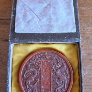 Op de afbeelding: Een ronde, roodbruine zegel met ingewikkelde draaksculpturen en Chinese karakters, gepresenteerd in een rechthoekige doos met een gele satijnen voering. De doos heeft een wit stoffen interieur en een klein handvat.