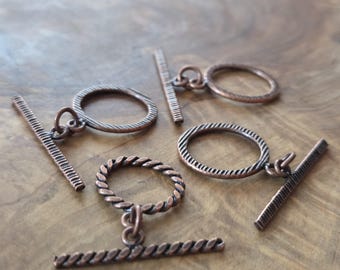 May include: Close-up of several copper-colored toggle clasps. The clasps feature a bar and ring design, with some rings having a twisted rope texture. The clasps are arranged on a wooden surface, showcasing their intricate details.