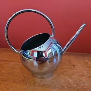 May include: A polished chrome watering can with a spherical body and a long, thin spout. It features a large, circular handle and a black interior. The surface reflects the surroundings, including a red wall and wooden table.