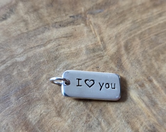May include: A small, rectangular silver charm with the words "I ♥ you" engraved in black. The charm has a small loop at one end for attaching to a chain or bracelet. The charm is displayed on a wooden surface.