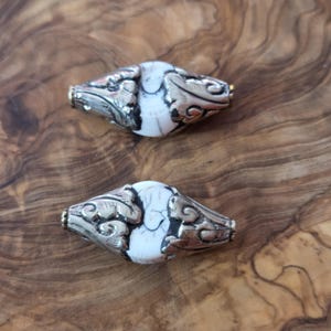 May include: Two decorative beads with a silver-colored metal design and a white and black stone center. The beads have a barrel shape with ornate metalwork and are displayed on a wooden surface.