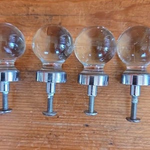 May include: Four clear glass ball cabinet knobs with silver-colored metal bases and threaded screws. The knobs are arranged in a row on a wooden surface. The glass spheres reflect light, creating a bright appearance.